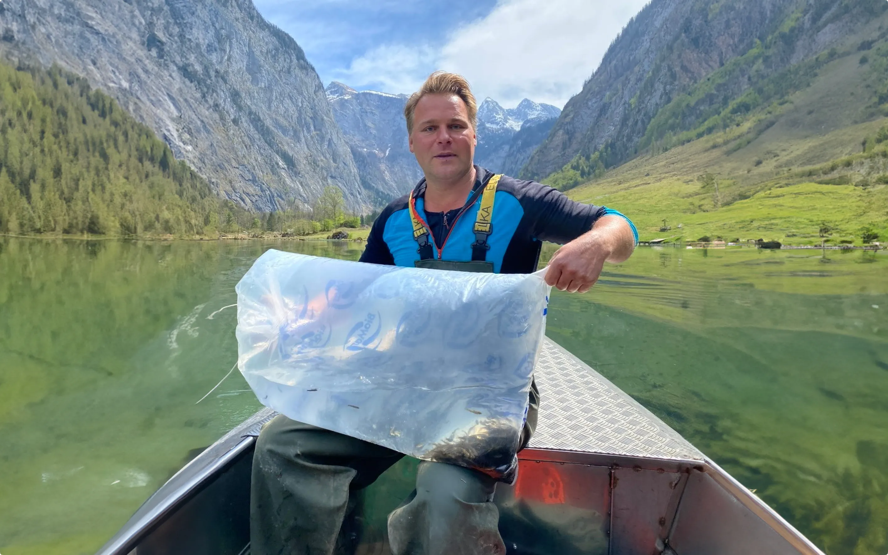 Ein Mann sitzt in einem kleinen Boot und hält einen großen, mit Wasser und Fischen gefüllten, Plastikbeutel. Das Boot schwimmt auf einem See. Im Hintergrund sind Berge zu sehen.